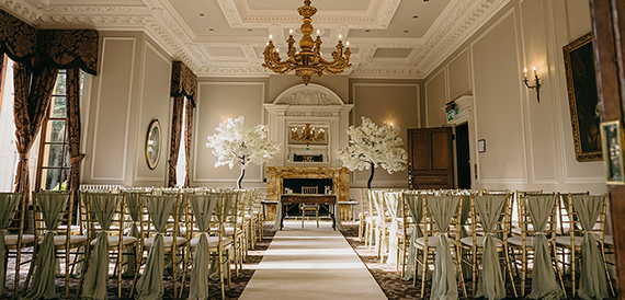 Wide view of a ceremony room with rows of sage green chair sashes leading to the fireplace