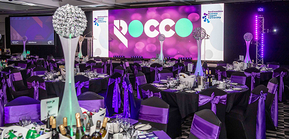 Gala dinner tables with silver centrepieces facing a pink-lit awards stage.