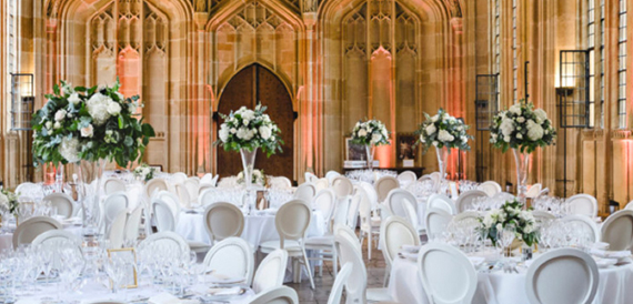 Tall white floral displays among round tables in a vaulted stone hall