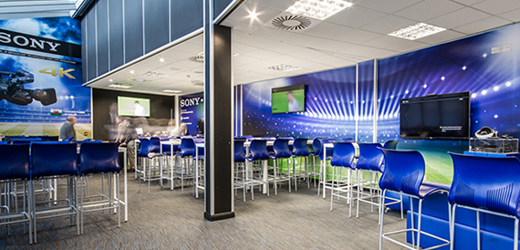 Sponsor lounge with blue bar stools and wall-mounted screens