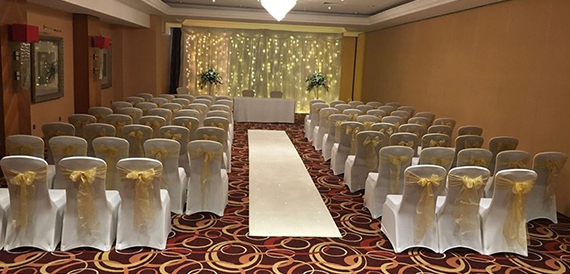 Wedding ceremony aisle with white chair covers, gold sashes and a fairy light backdrop
