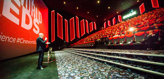 Cinema auditorium with tiered red seating and a presenter on stage