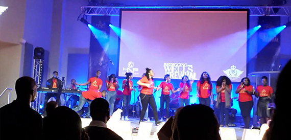 Performers in red shirts singing on stage before a Wells Revival screen backdrop