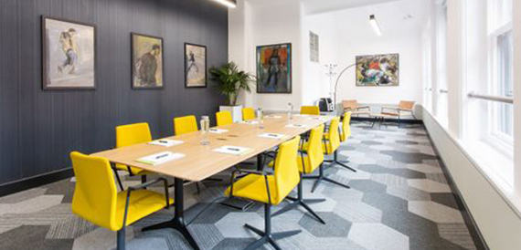 Long meeting table lined with yellow chairs beneath framed artwork
