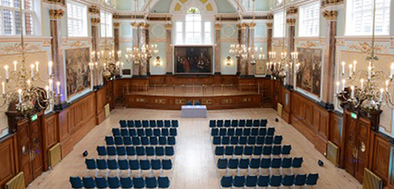 Wood-panelled hall set theatre-style with rows of blue chairs facing the stage