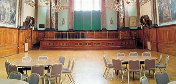Wood-panelled hall with round tables set boardroom-style before green screen panels