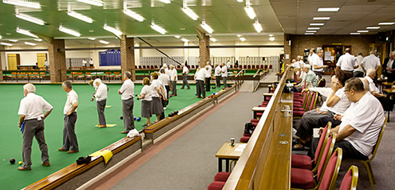 Indoor bowls green with players in whites and spectators seated in red chairs