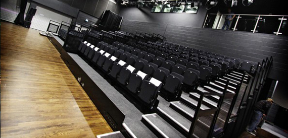 Tiered auditorium with rows of black fold-down seats facing the stage.