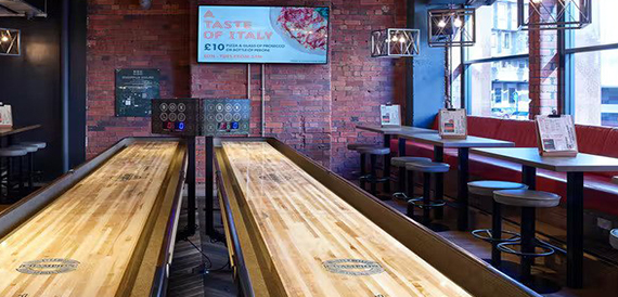 Shuffleboard tables beside red booth seating and a wall screen