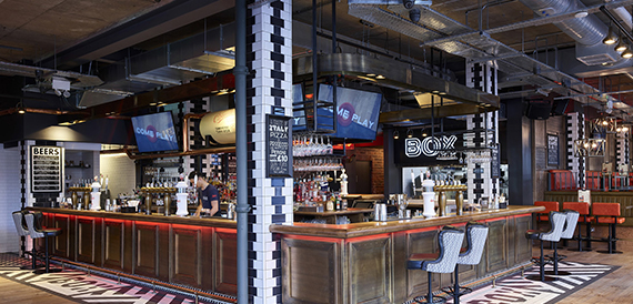 Bar counter serving drinks below screens and a chequered pillar