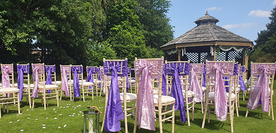 Outdoor wedding ceremony chairs with purple sashes facing a garden gazebo.