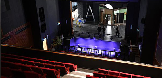 View from a theatre balcony over red seating towards a lit backstage area.