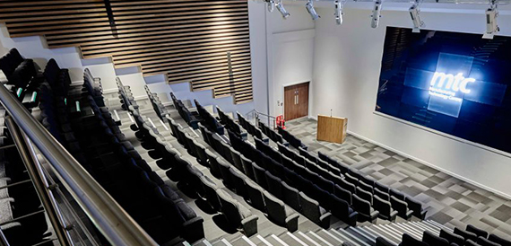 Tiered auditorium with black seating facing a lectern and screen showing the MTC logo.