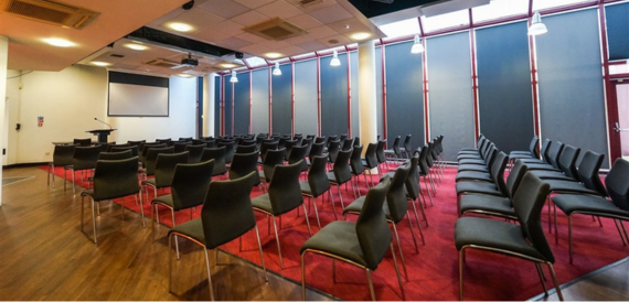 Conference room set theatre-style with dark chairs on a red carpet facing a lectern