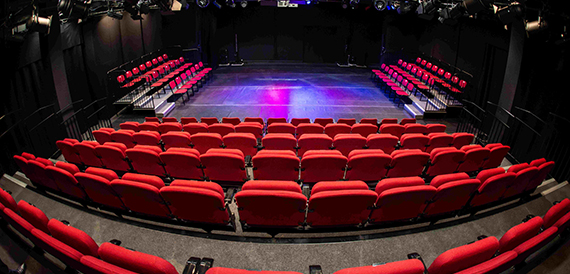 Theatre auditorium with red tiered seating facing an empty stage lit purple and blue.