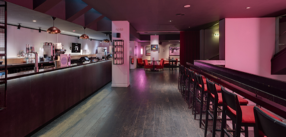 Bar counter with hanging lamps and red stools along a pink-lit corridor