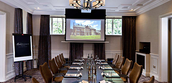 Boardroom with a long green-clothed table, leather chairs and a screen showing the hall's exterior