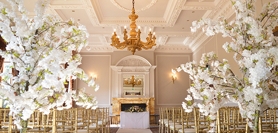 Ceremony room with gold chairs, an ornate ceiling and blossom trees either side of the fireplace