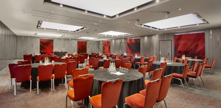 Banqueting room with round tables, orange velvet chairs and red abstract art panels on grey walls.