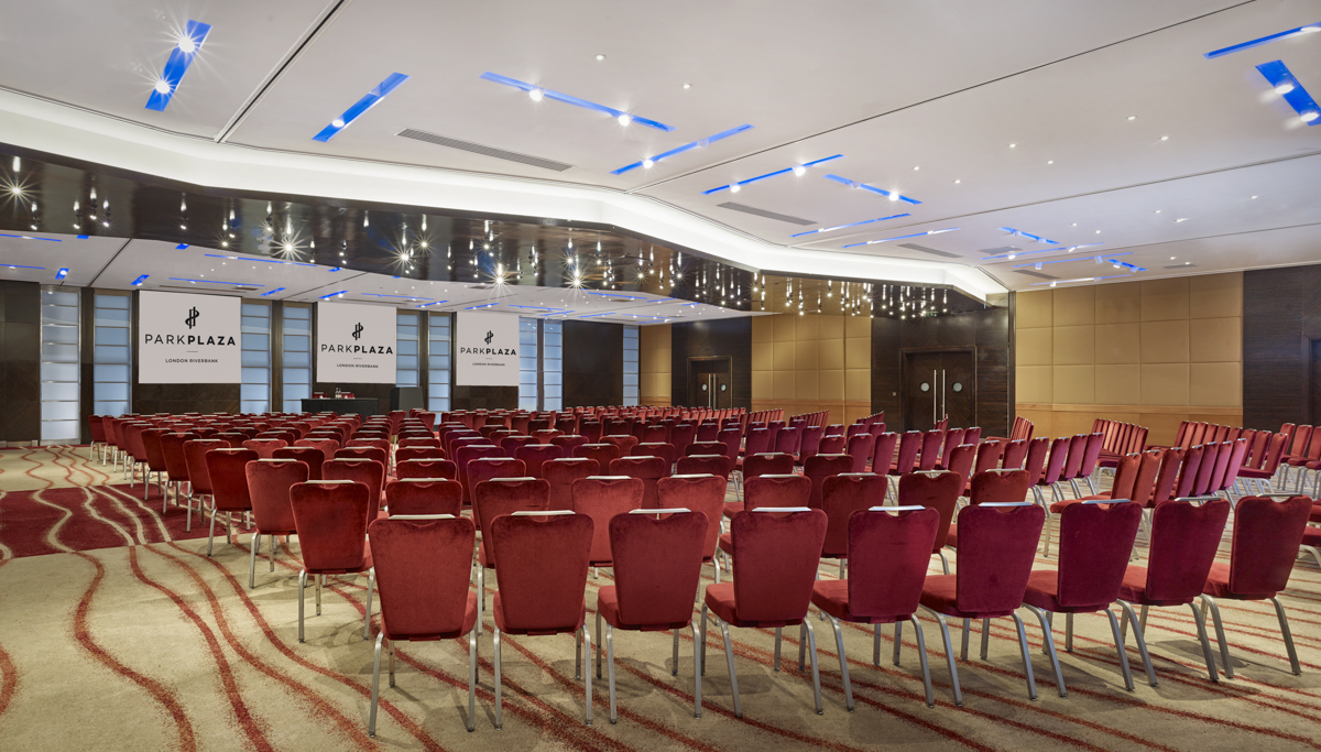 Conference hall with rows of red velvet chairs facing a stage with Park Plaza Riverbank signage.