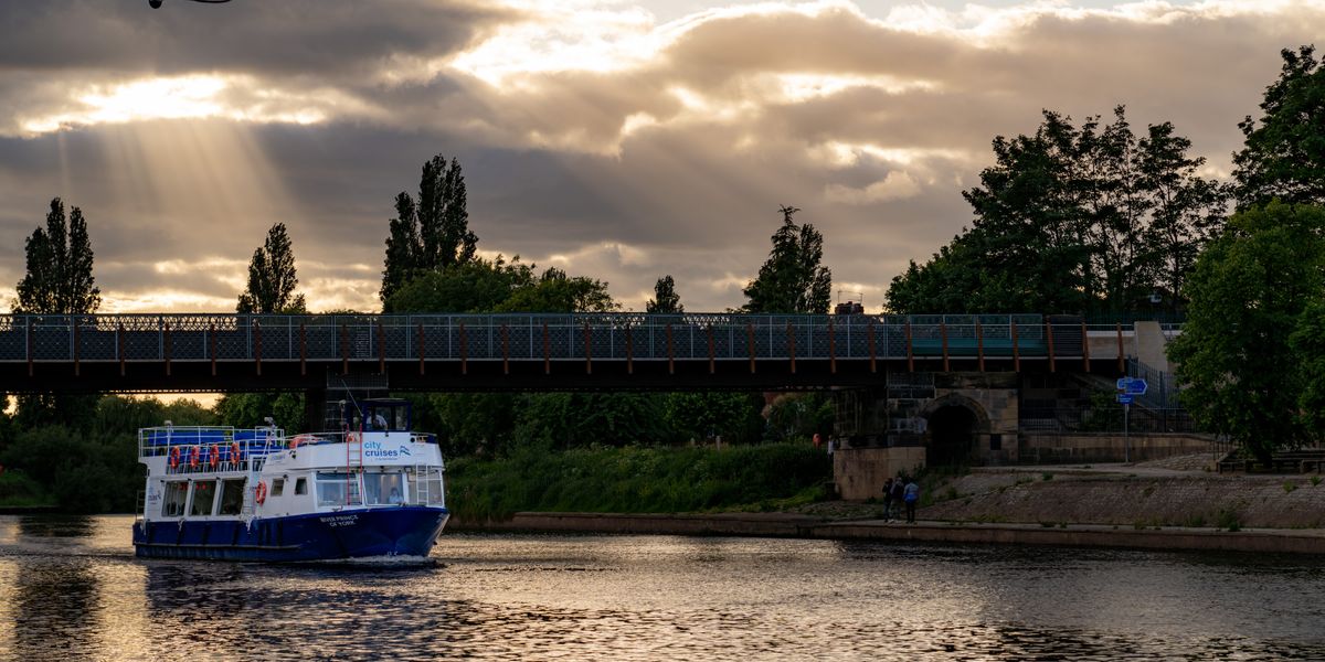 River Prince boat cruising beneath a railway bridge at dusk with sun rays through clouds