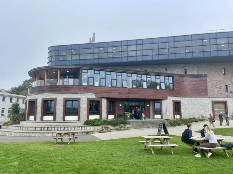 Campus building exterior with picnic tables on the lawn and students seated outside.