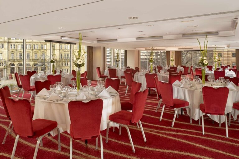 Banqueting room with round tables, white linen, flower arrangements and red velvet chairs.